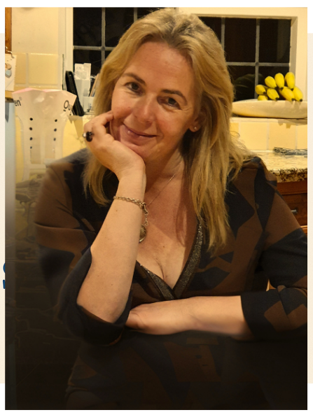 Sleep practitioner Jane A woman smiling, wrapped in a large grey blanket, sitting at a table in a café.