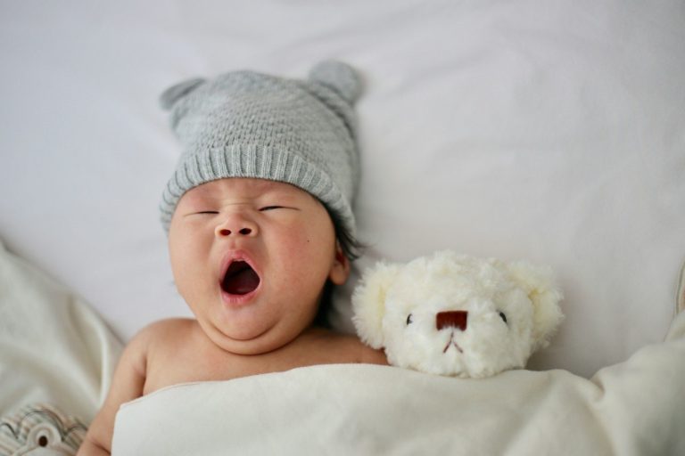 Sleeping baby Baby wearing a grey hat yawning, lying beside a toy bear on a soft blanket.